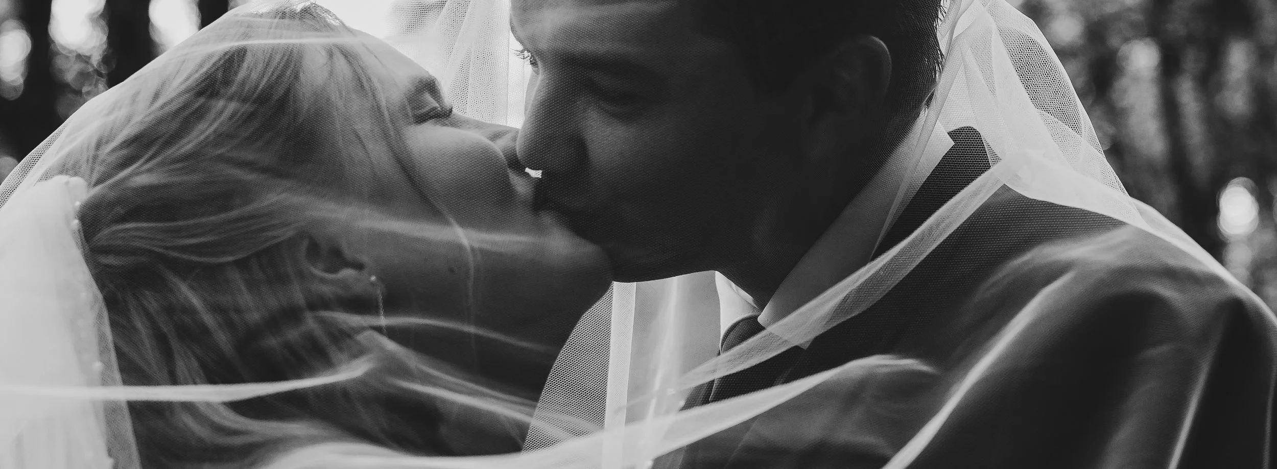 A black and white photo of a couple sharing a kiss, with the bride wearing a veil and earrings, and the groom in a suit. They are close together outdoors, with blurred trees in the background.