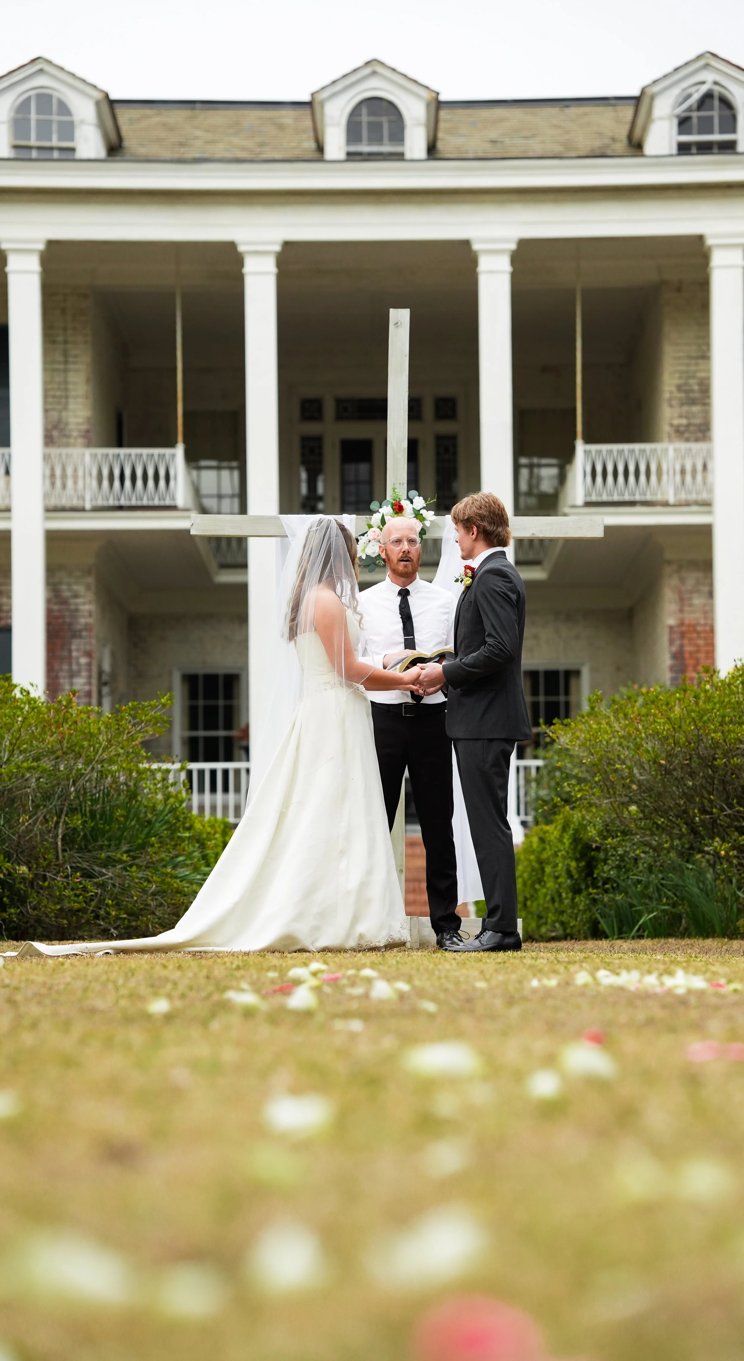 A couple dressed in wedding attire standing in front of a white cross decorated with flowers during an outdoor wedding ceremony in front of a large house.