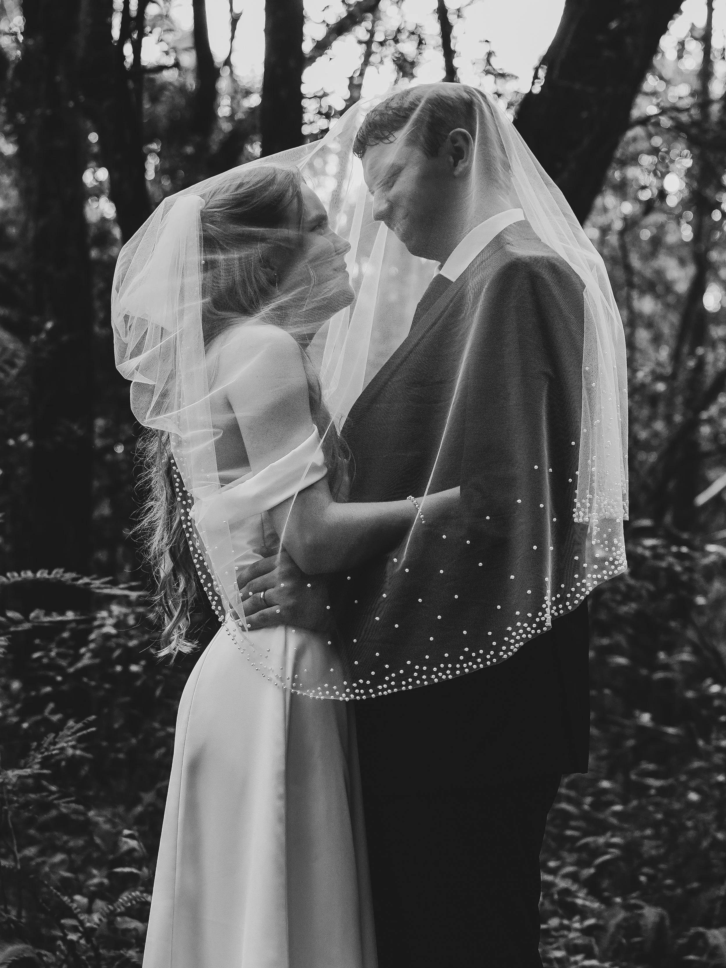 A black and white photo of a bride and groom standing close together outdoors, wrapped in a sheer veil, with trees in the background.