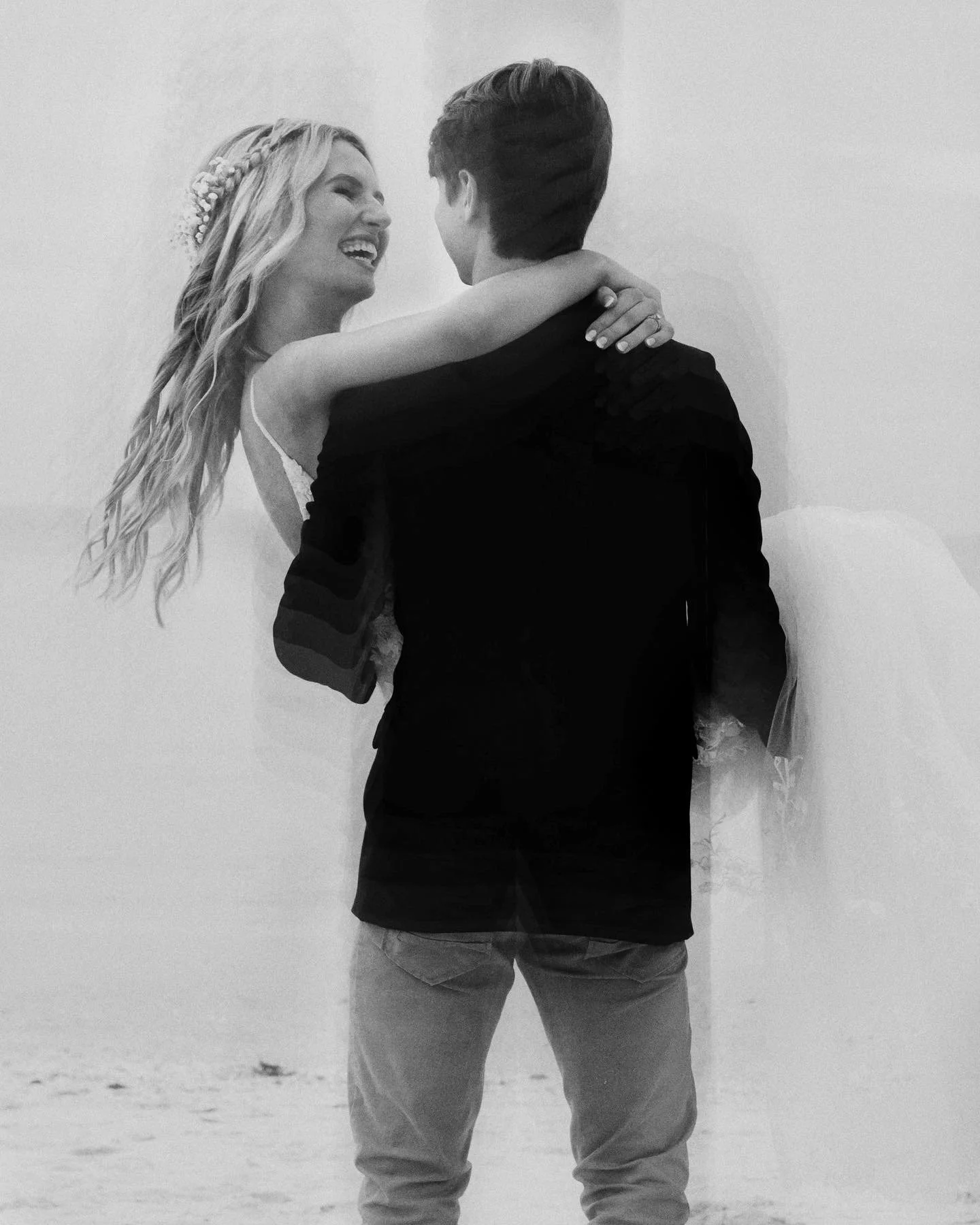 A black and white photo of a couple dancing on the beach, with the woman smiling and looking at the man, who is holding her. The woman has long, wavy hair and the man has short hair. The background shows sand and a cloudy sky.