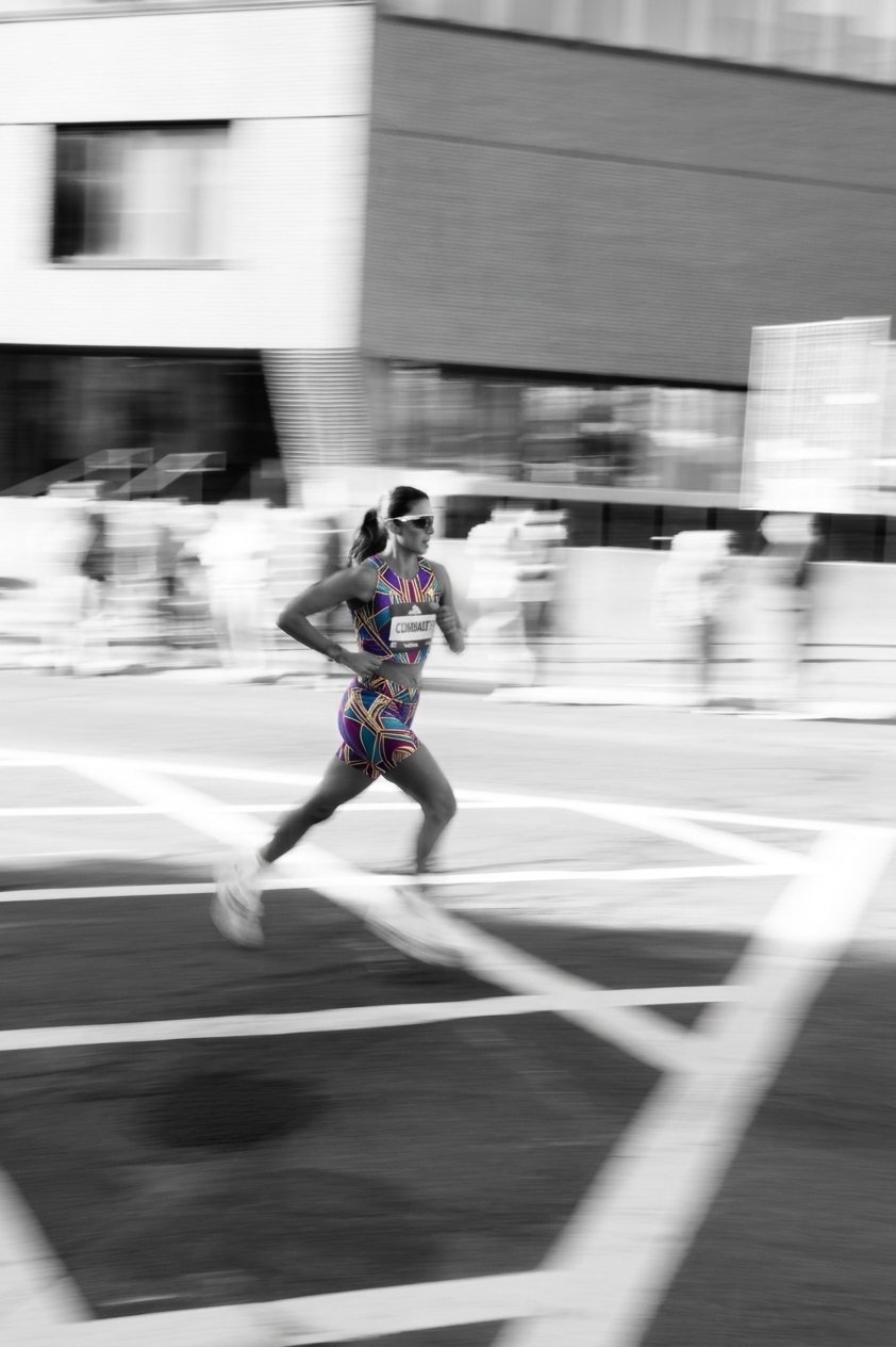 Female runner wearing sunglasses and a colorful athletic outfit races through a city street in a motion-blurred black-and-white scene.