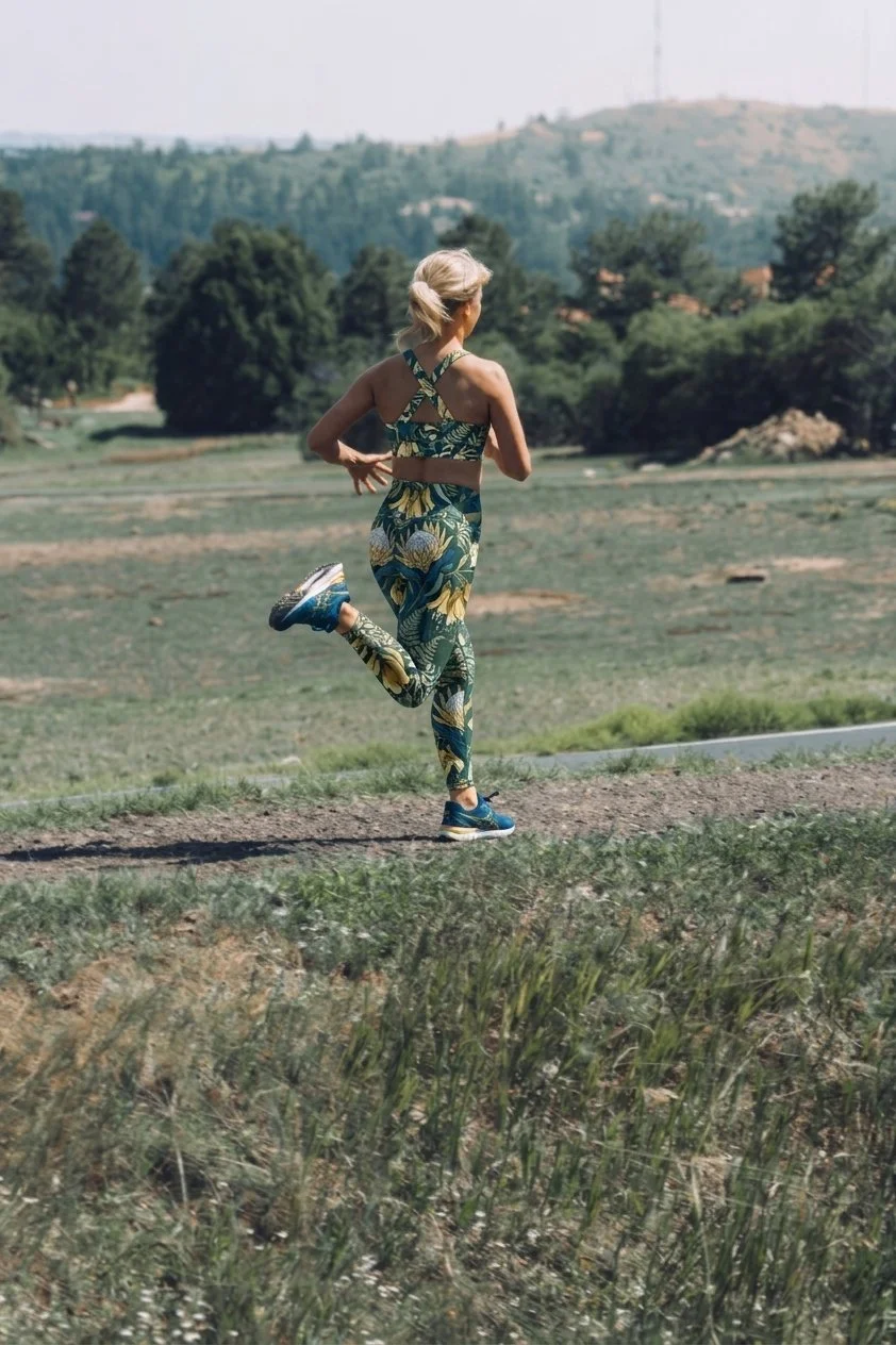 Woman jogging outdoors on a dirt trail in a patterned athletic outfit, with trees and hills in the background