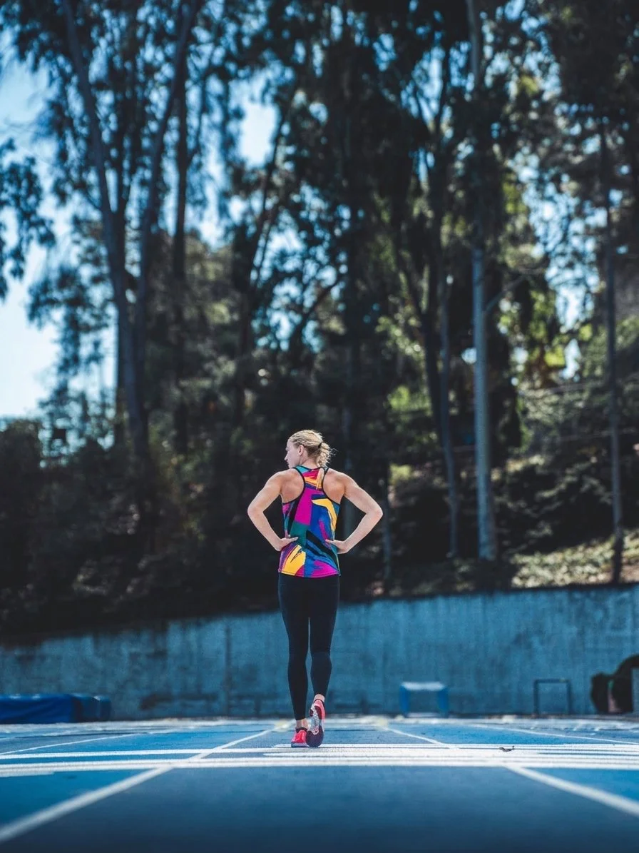 Female runner standing on an outdoor track in a colorful athletic tank top and black leggings, surrounded by trees