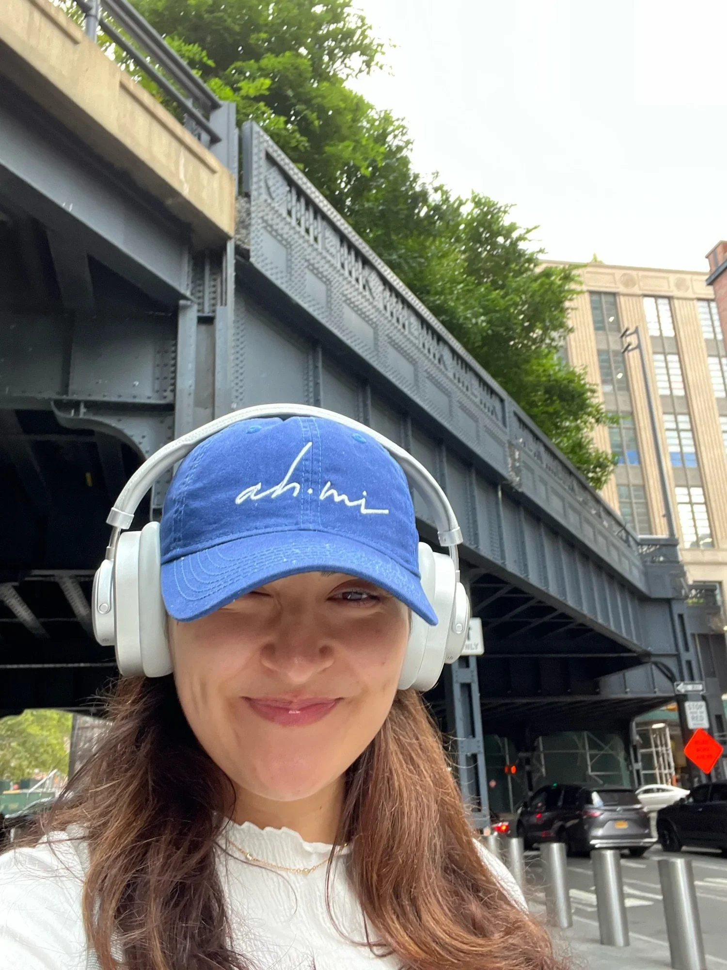 Selfie of a woman wearing a blue baseball cap and white headphones beneath a steel bridge in an urban street.