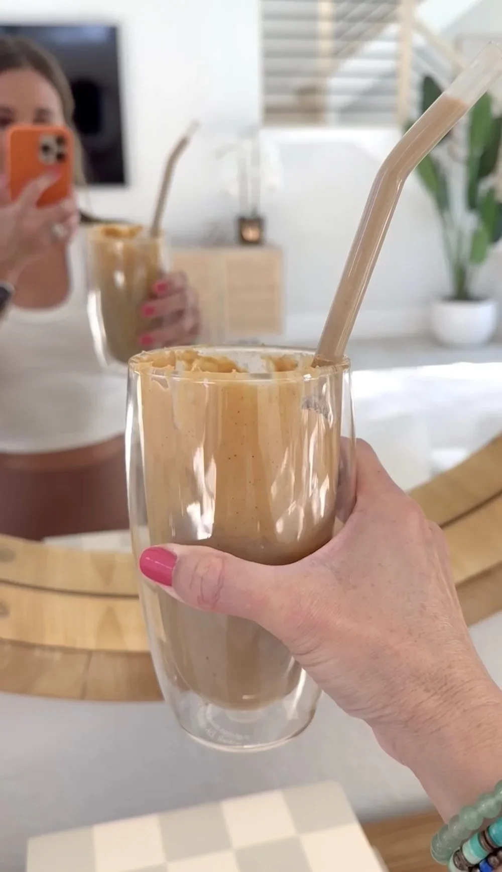 Hand holding a glass of creamy iced coffee with a reusable straw, reflected beside another drink