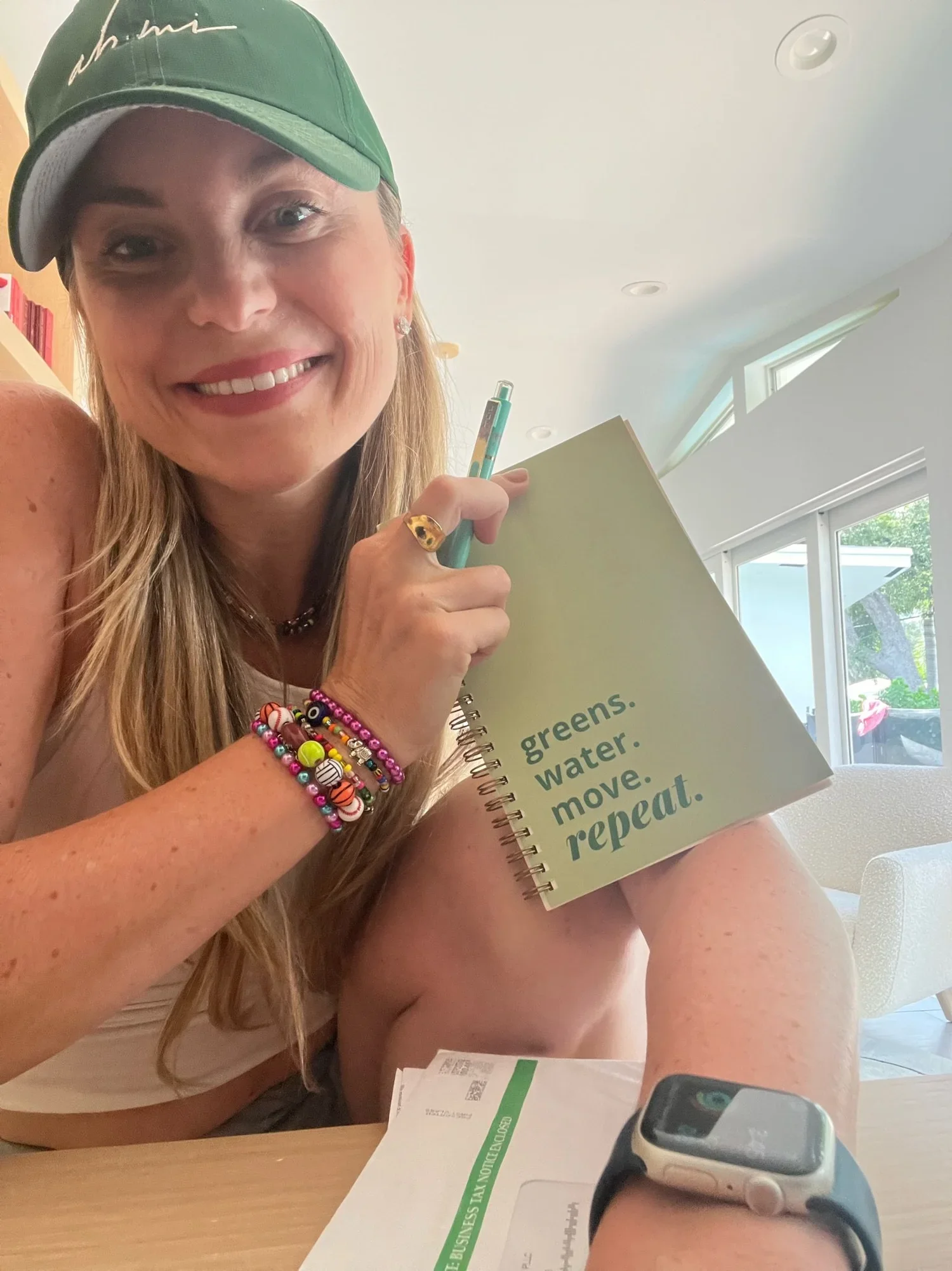 Smiling woman in a green cap holding a notebook and pen at a sunny desk