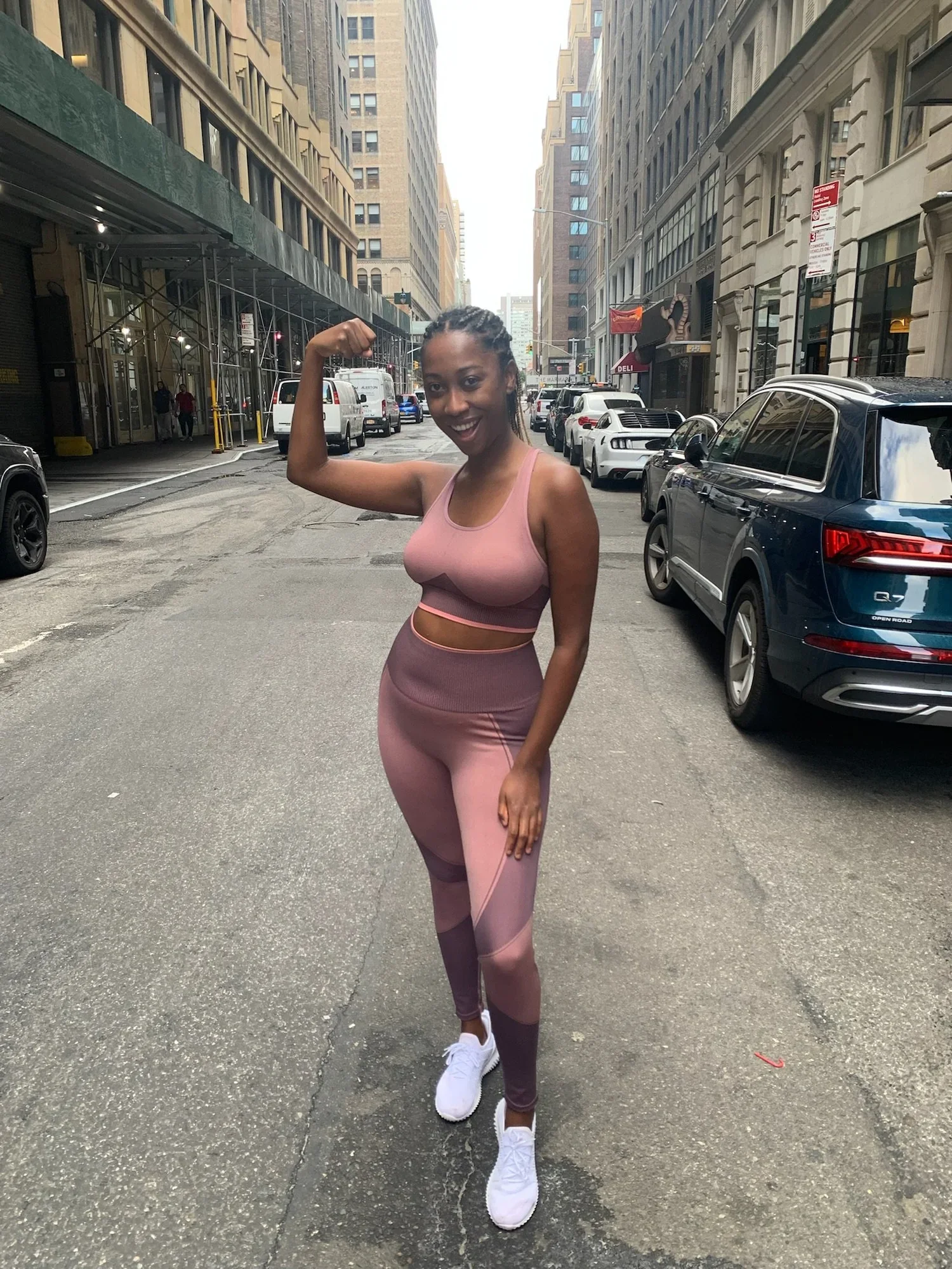 Woman in pink athletic wear flexing on a downtown city street surrounded by tall buildings and parked cars