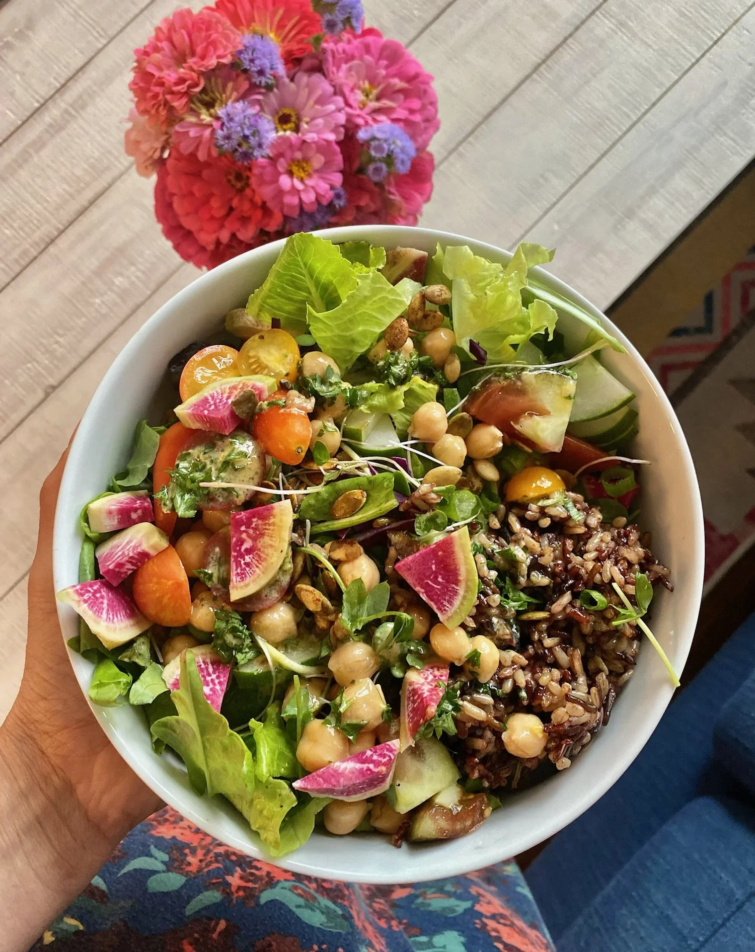 Colorful grain and chickpea salad with greens, tomatoes, cucumber, watermelon radish, seeds, and microgreens, with a bouquet of flowers in the background
