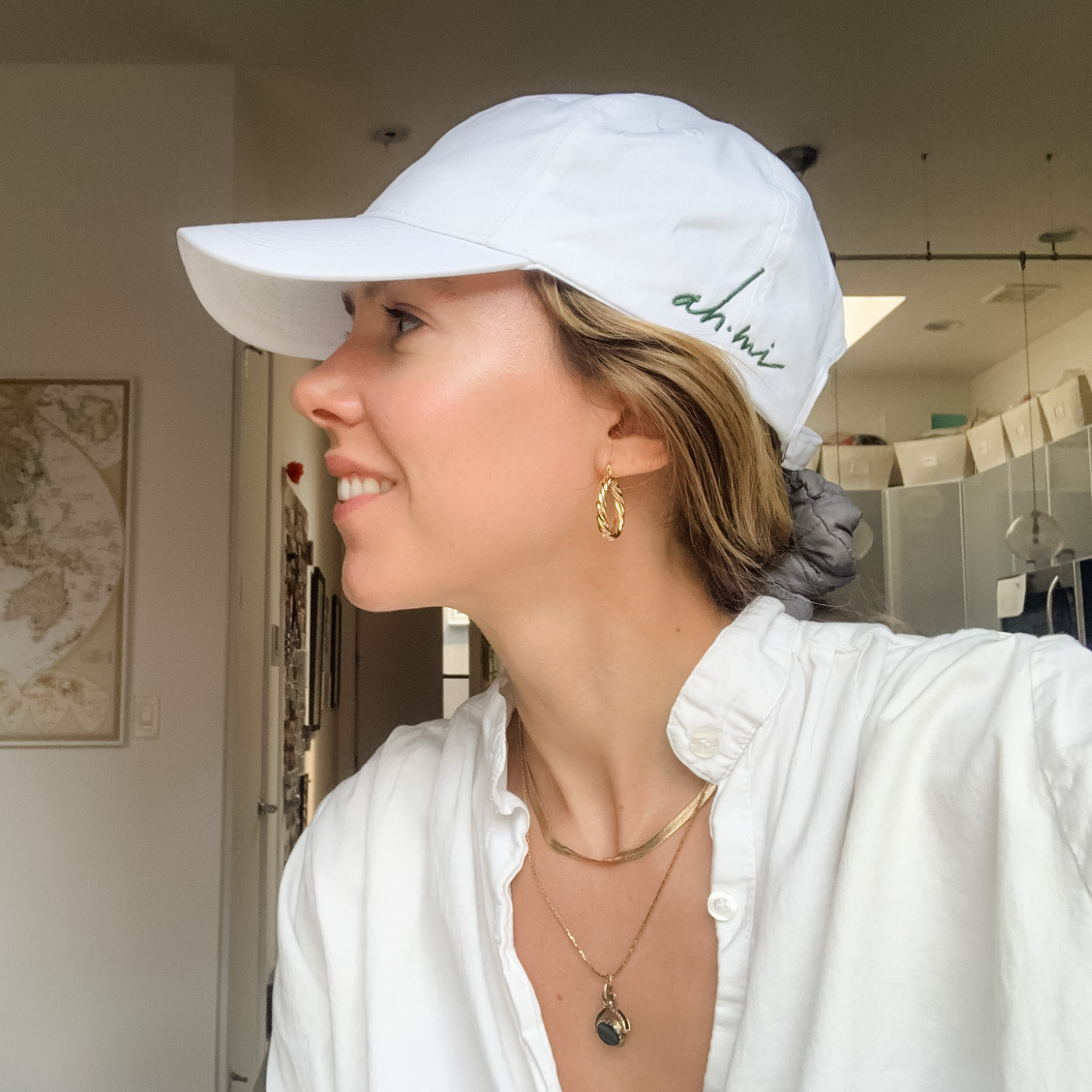 Smiling woman in profile wearing a white baseball cap, white shirt, gold hoop earrings, and layered necklaces indoors