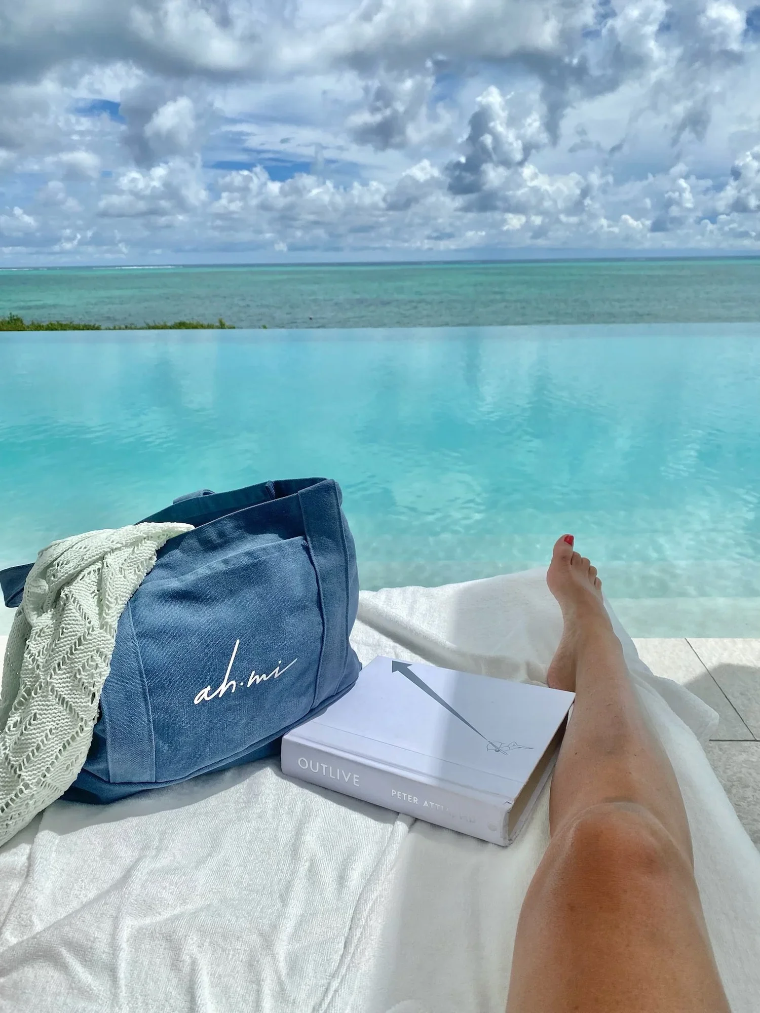 Poolside view of turquoise ocean water, a blue beach tote, white towel, book, and reclining person under a cloudy sky