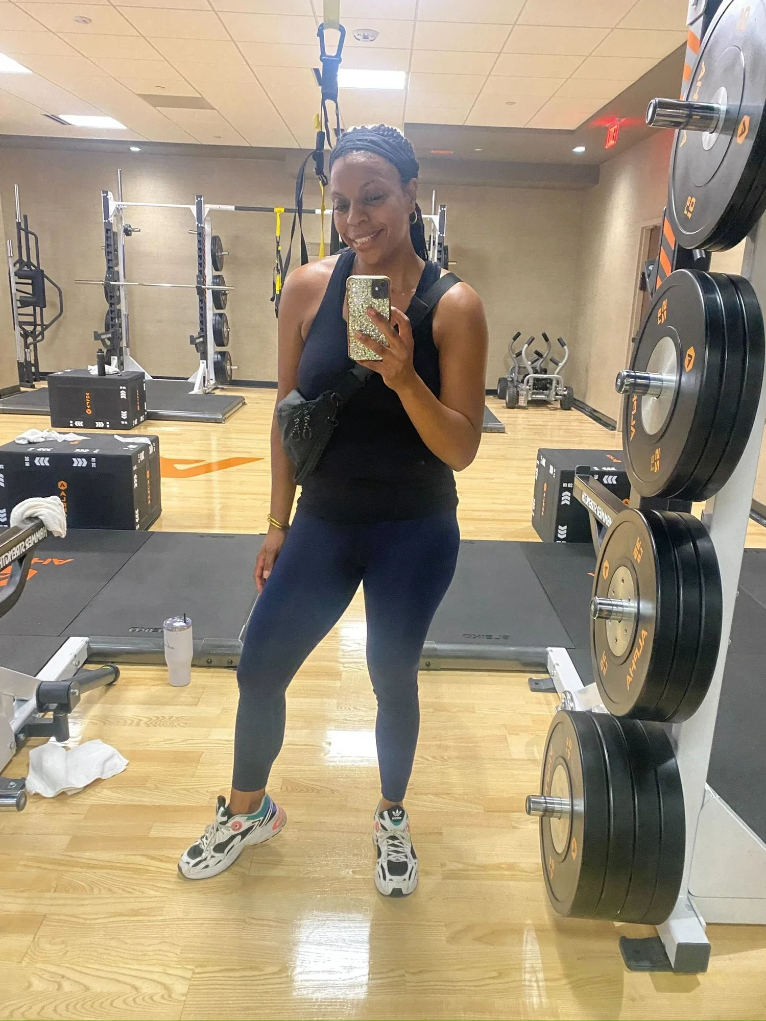 Woman taking a mirror selfie in a gym, wearing black workout clothes and standing beside weightlifting equipment.
