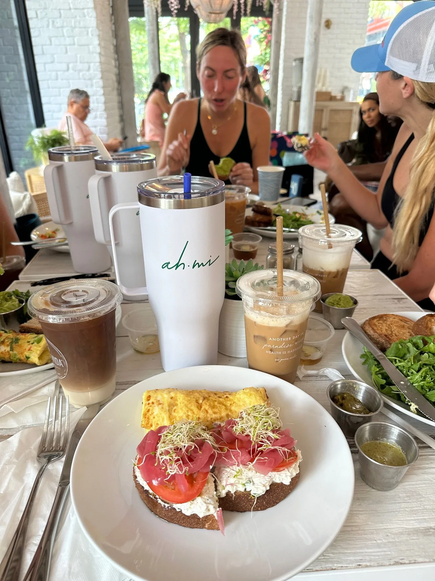 Friends dining at a bright cafe with avocado toast, omelet, salads, iced coffee, and branded tumblers on the table.