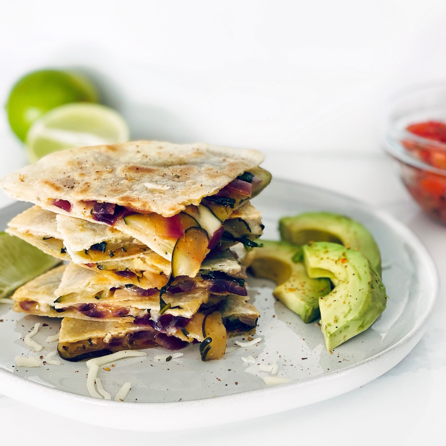 Stack of vegetable quesadillas with melted cheese, avocado slices, lime wedges, and salsa