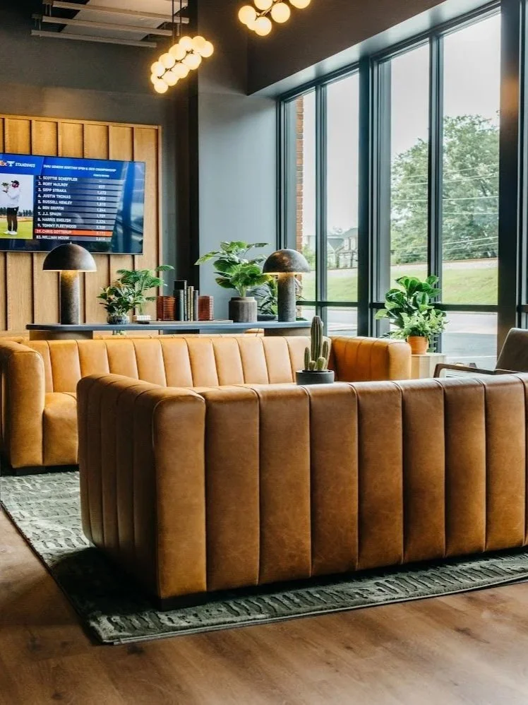 Modern lounge area with leather sofas, large windows, plants, and a TV on a wooden accent wall.