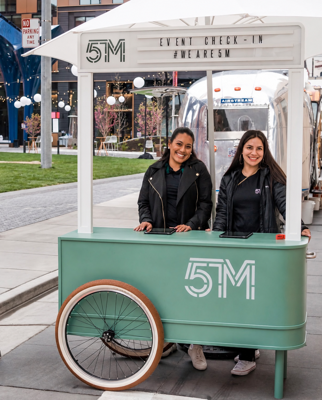 Two women standing inside a teal-colored mobile cart with a large wheel. The cart displays event check-in signage and the hashtag #WEARE5M. The women are smiling, wearing black jackets, and appear to be at an outdoor event.
