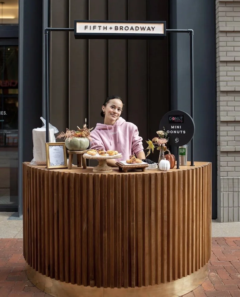 A woman standing behind a wooden circular booth with baked goods and decorative pumpkins, under a sign that reads 'FIFTH + BROADWAY'.