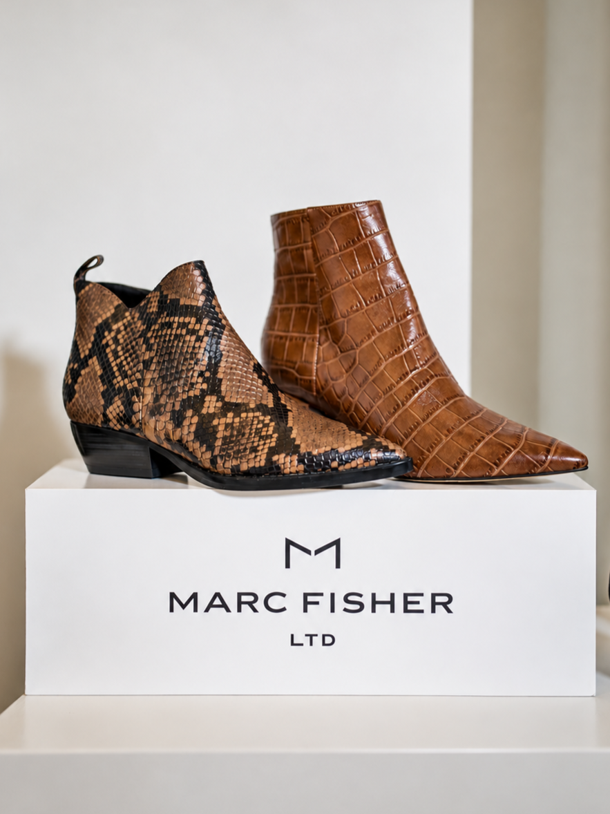 A pair of designer boots, one with a snakeskin pattern and the other with a crocodile texture, displayed on a white box with the Marc Fisher logo.