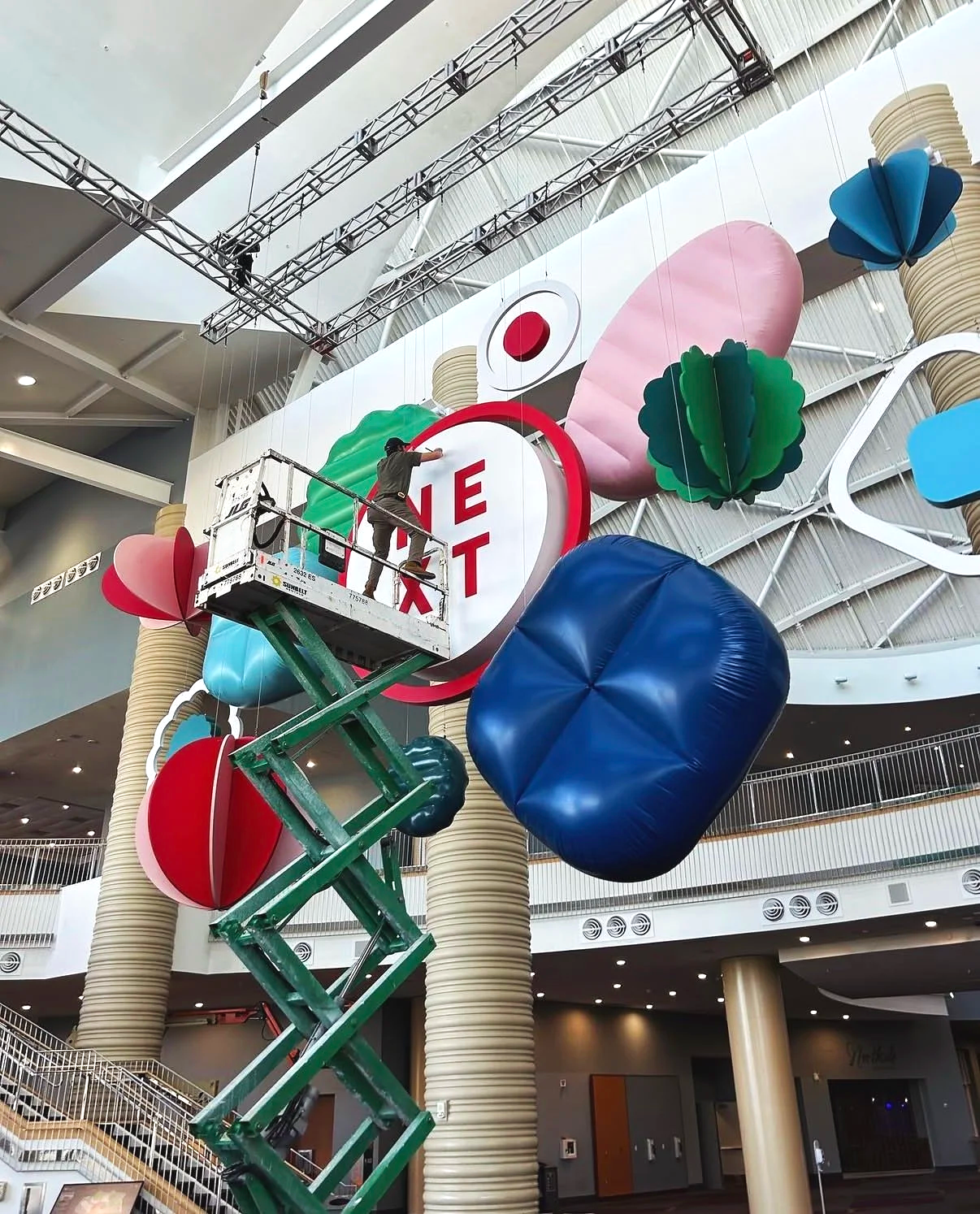 A worker on a lift installing a large Coca-Cola sign in a shopping mall, with colorful oversized inflatables of bottles, berries, and a lemon in the background.