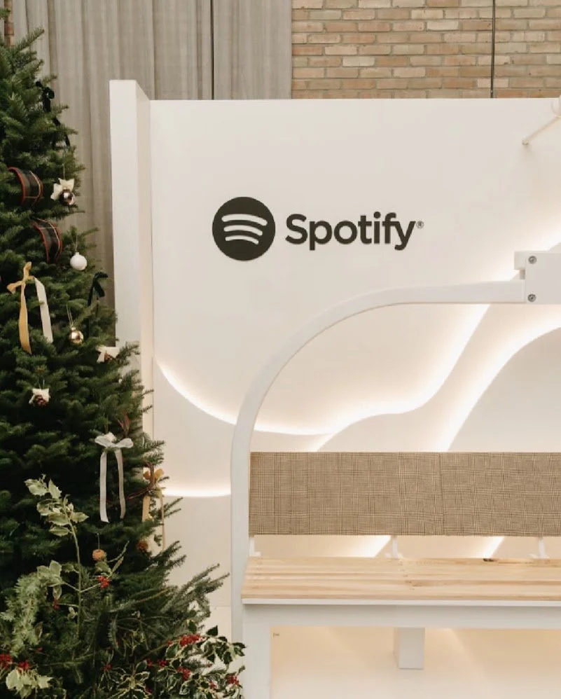 A Christmas tree decorated with ornaments and ribbons next to a white Spotify booth with a bench, set against a brick wall.