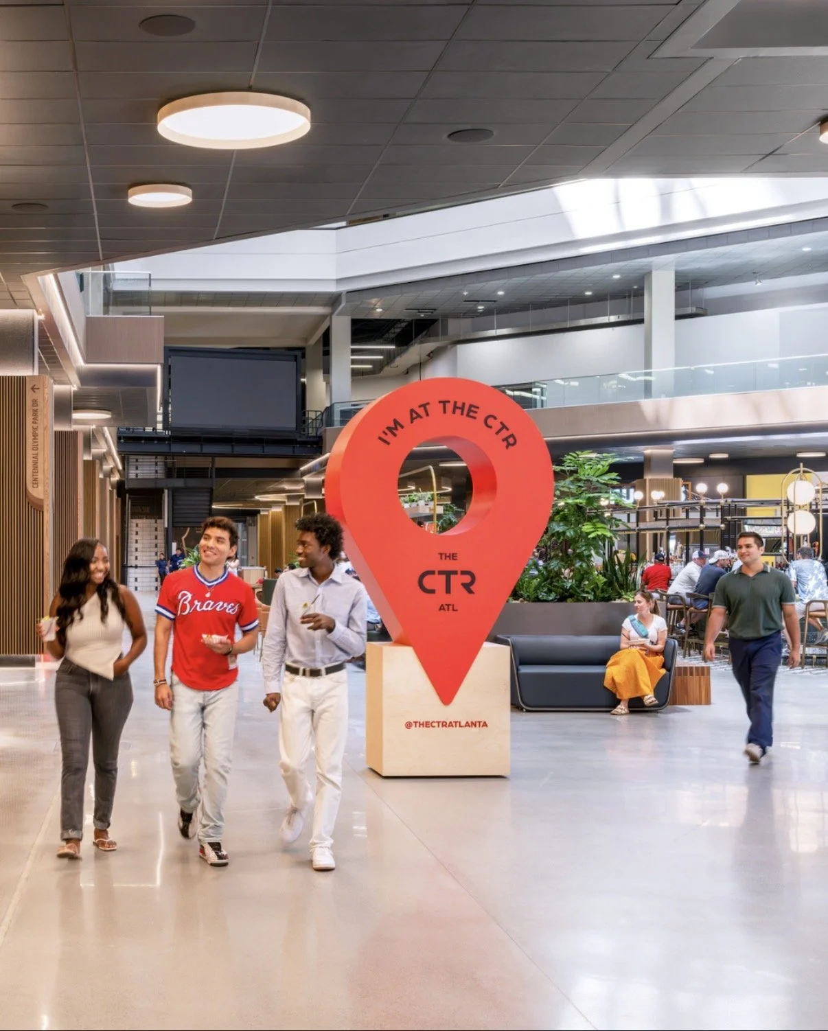 People walking inside a modern shopping mall, with a large red map pin sculpture that says "I'm at the CTR ATL."