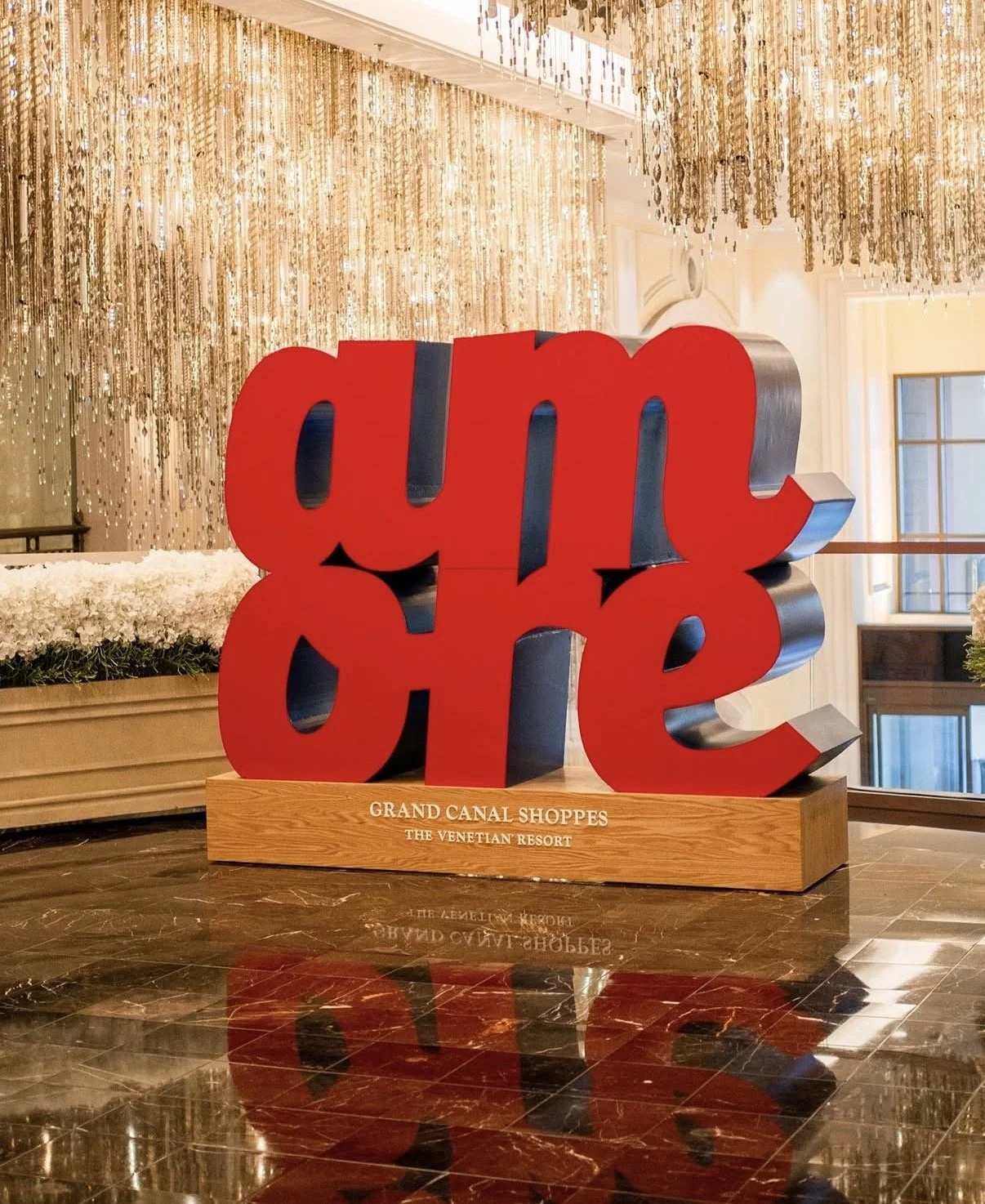 Large red and black sculpture spelling "yum cha" on a wooden base with white flowers and a chandelier in the background at the Grand Canal Shoppes, The Venetian Resort.