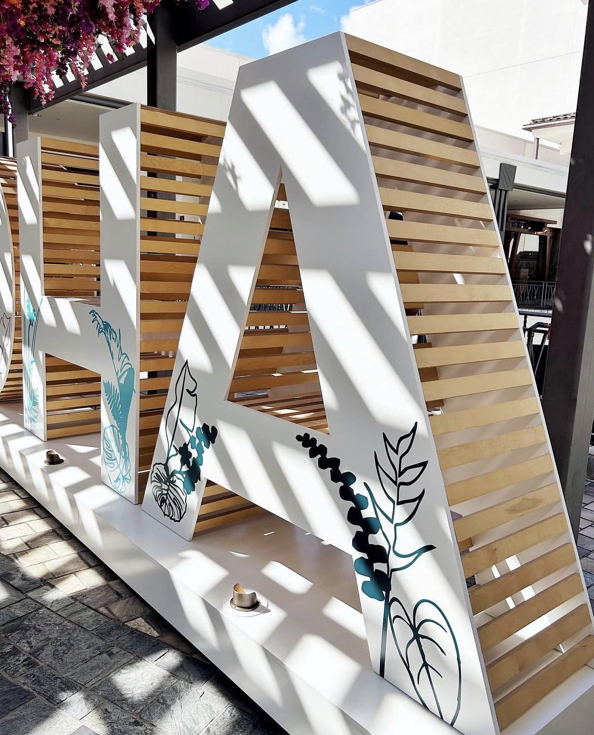 Decorative outdoor installation with large white letter 'A' featuring botanical illustrations, casting shadows on a paved surface.