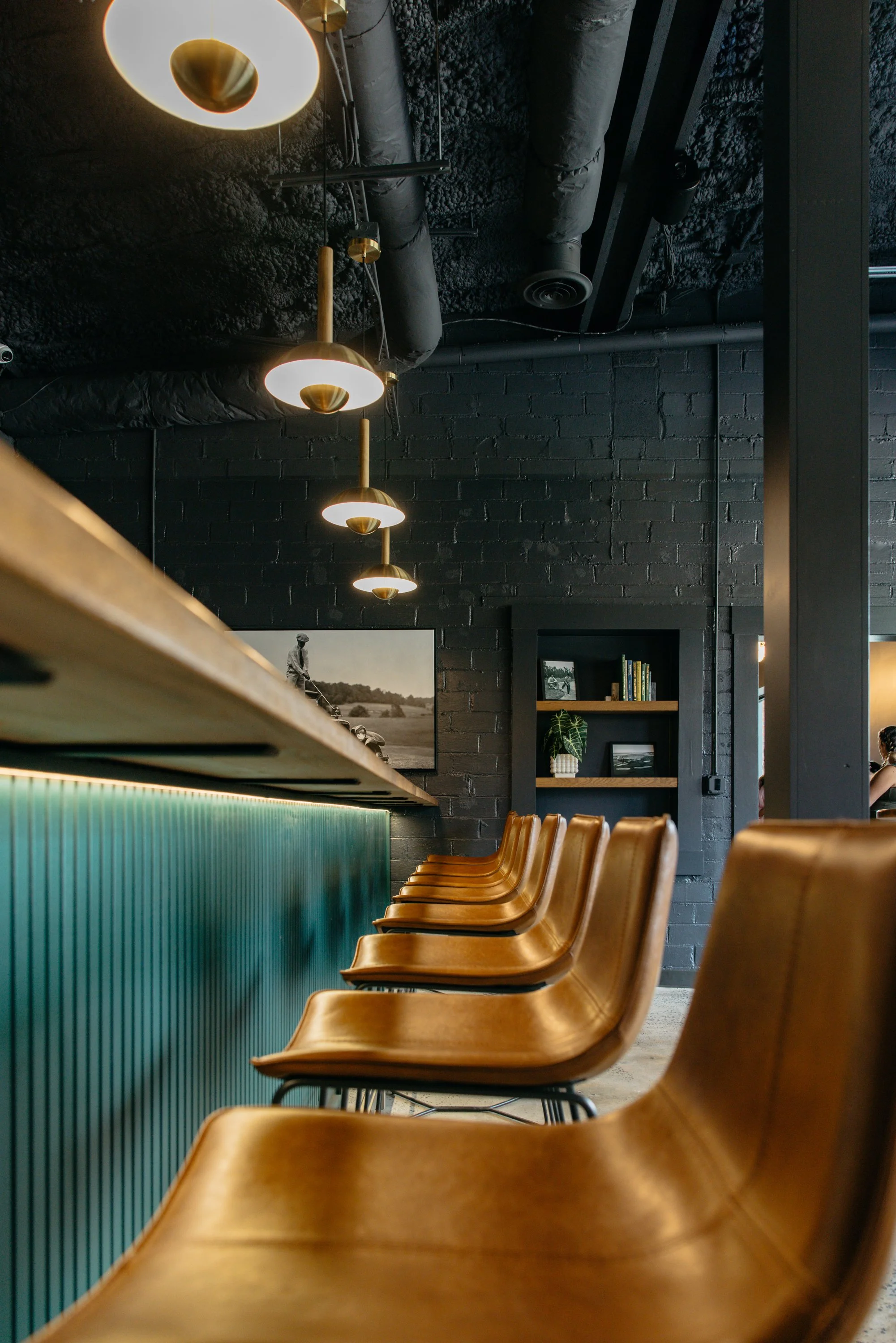 Interior of a modern café or restaurant with a black brick wall, row of wooden chairs, contemporary lighting fixtures, and shelving with books and plants.