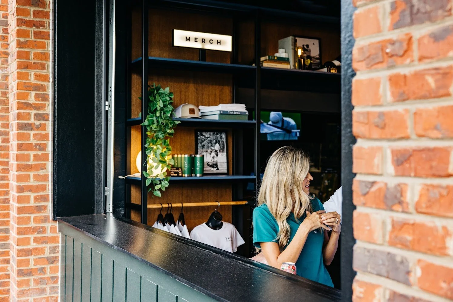 A woman with blonde hair in a teal shirt holding a cupcake at a counter behind a window in a brick-walled shop.