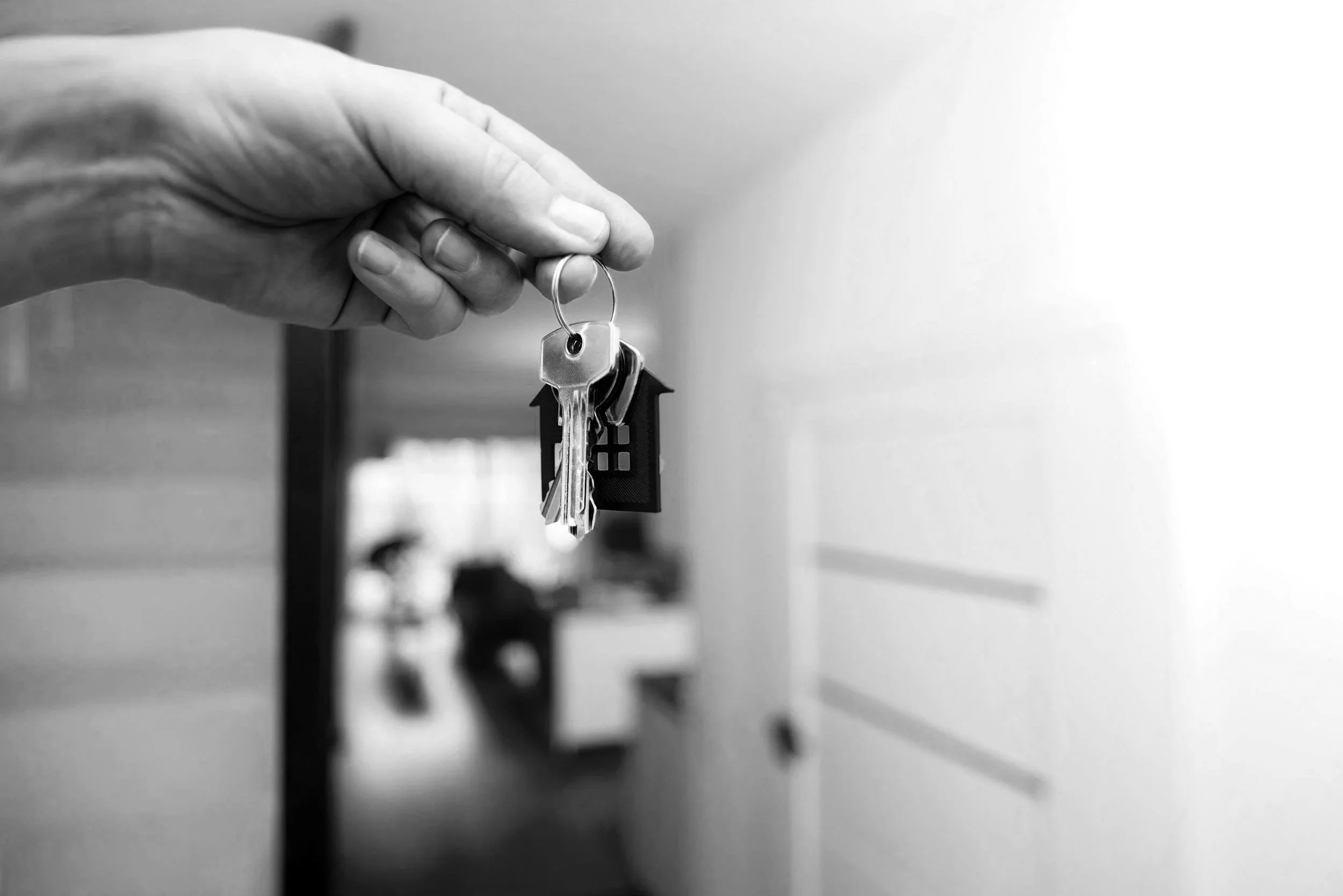 A hand holding a keychain with a small house-shaped keyring and keys, in black and white.