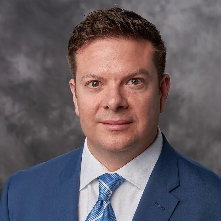 Professional headshot of a man in a blue suit, white shirt, and blue striped tie against a gray background.