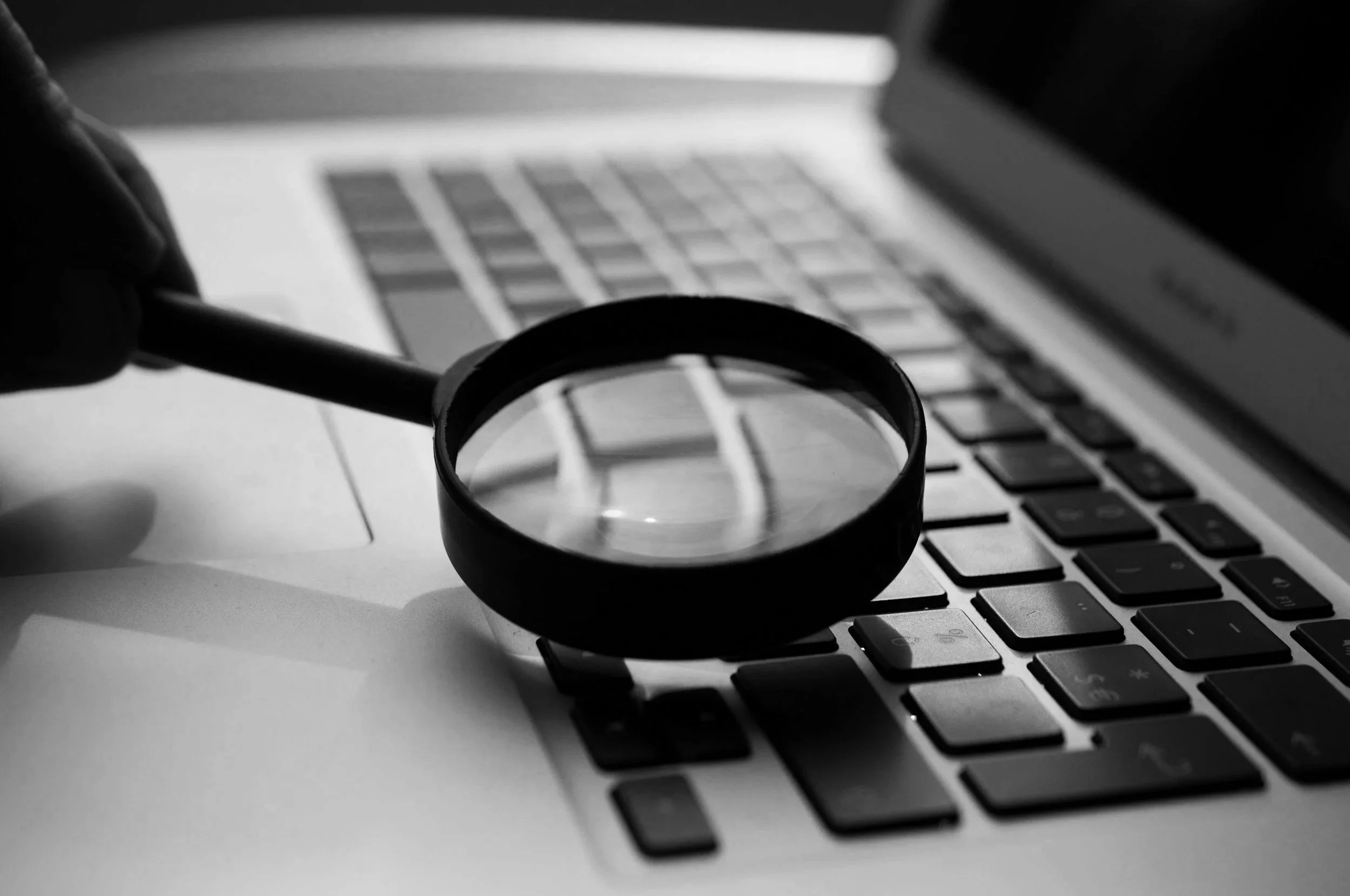 A magnifying glass held over a laptop keyboard with a blurred laptop screen in the background, in black and white.