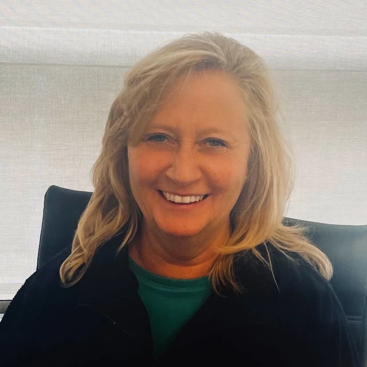 A woman with blonde hair smiling, sitting in an office chair in front of a white textured wall, wearing a black jacket and green shirt.