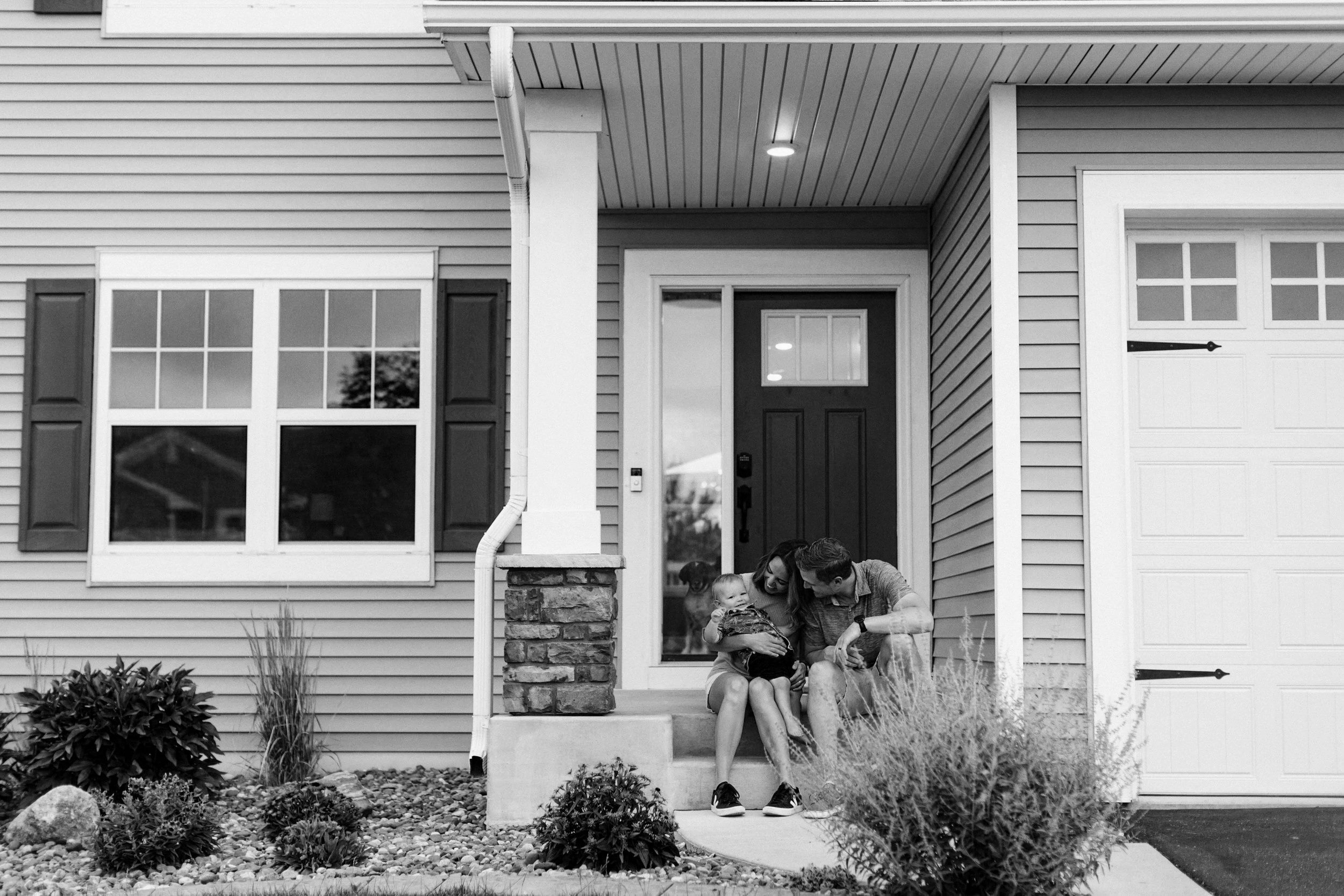 A family of three sitting on the front porch of a house, with a child and a dog, in black and white.