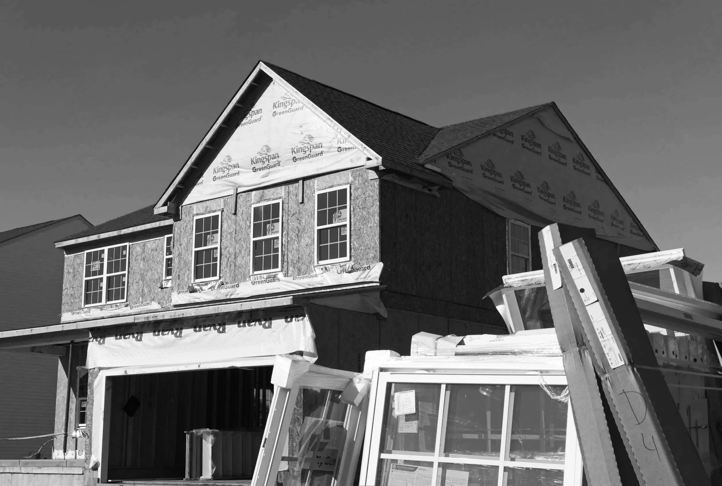 A house under construction with a partially finished exterior and windows. There are building materials and windows leaning against the house. The sky is clear.