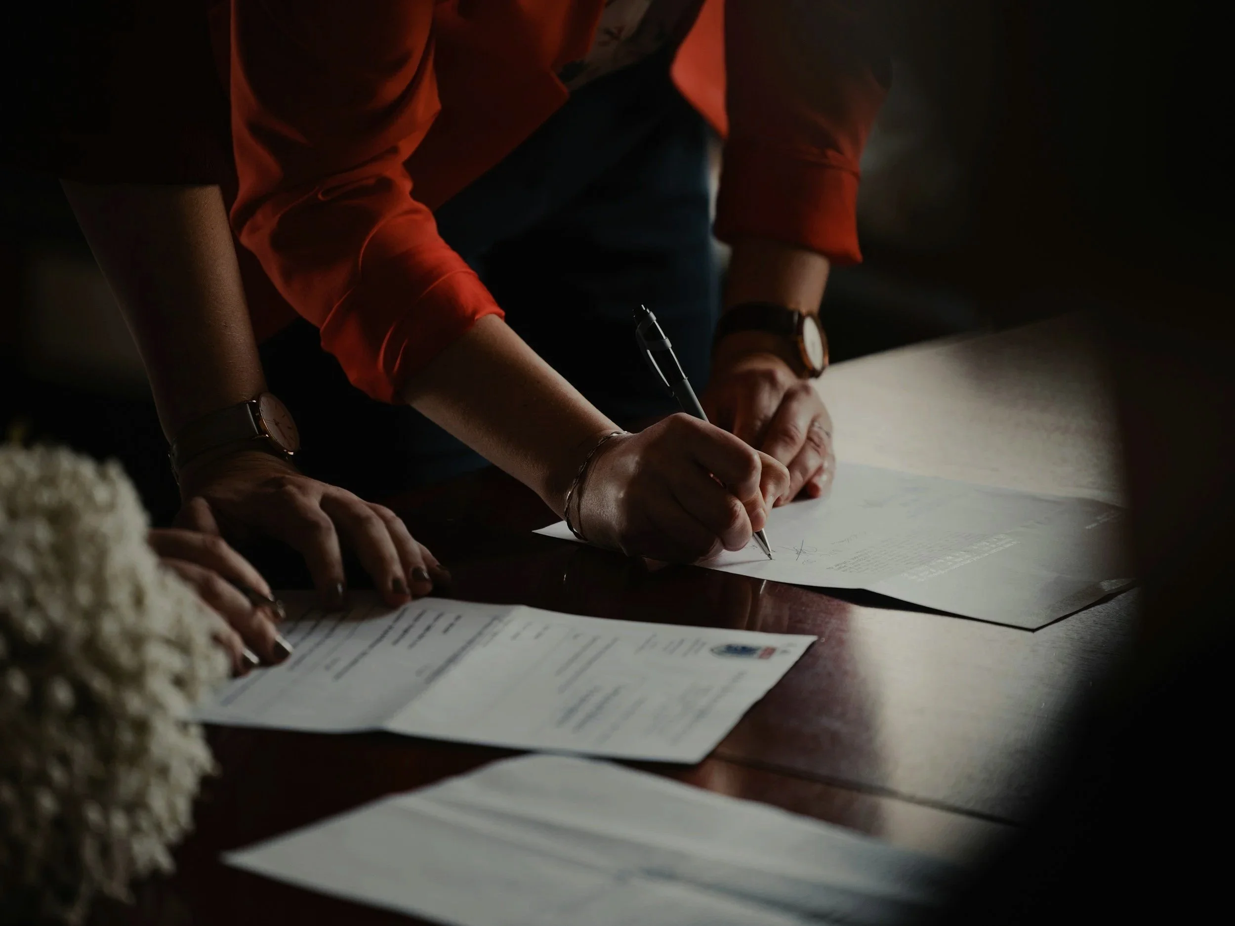 A person wearing an orange shirt and a watch is writing on a piece of paper on a wooden table, with other papers spread out on the table.