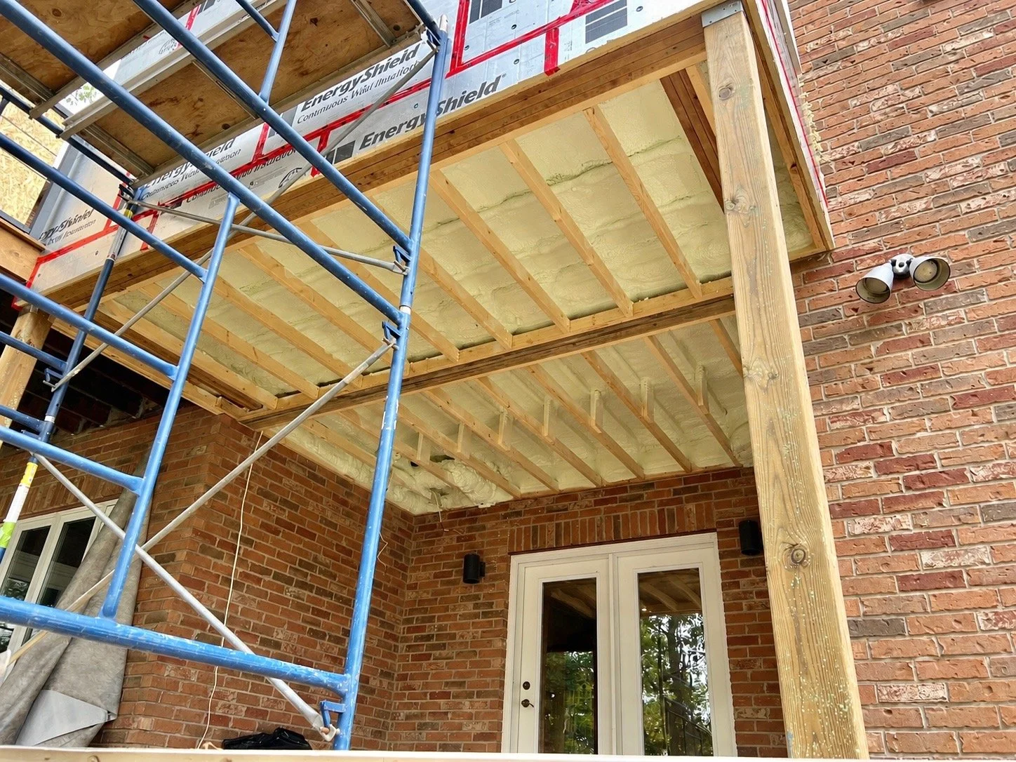 Construction site showing a partially built second-story deck with roofing insulation, wooden framing, scaffolding, brick exterior wall, and a double glass door leading inside