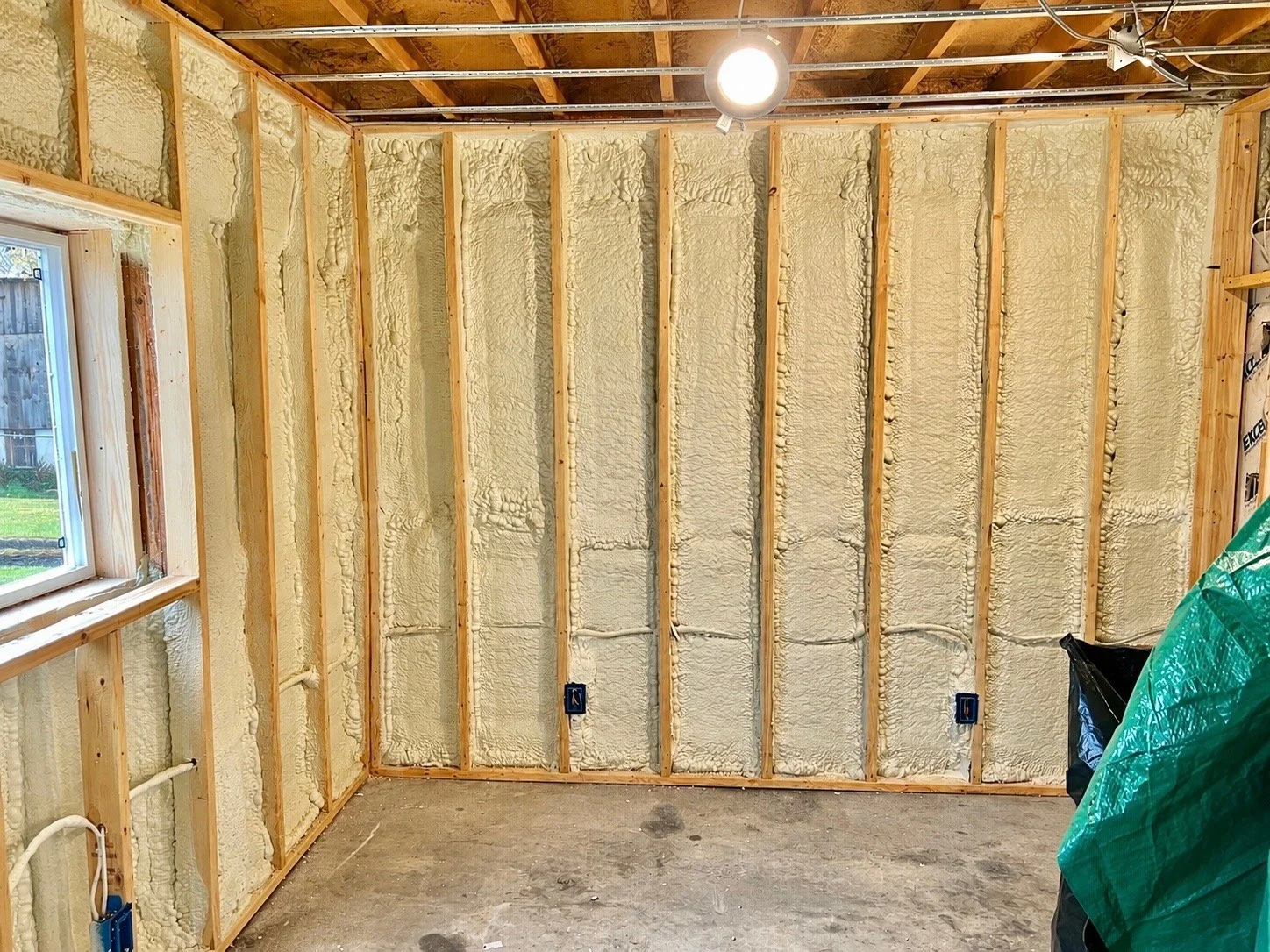 Interior of a room under construction with insulation installed in the walls, exposed ceiling beams, and electrical outlets. There's a window on the left side showing a view of a backyard with grass and a wooden fence.