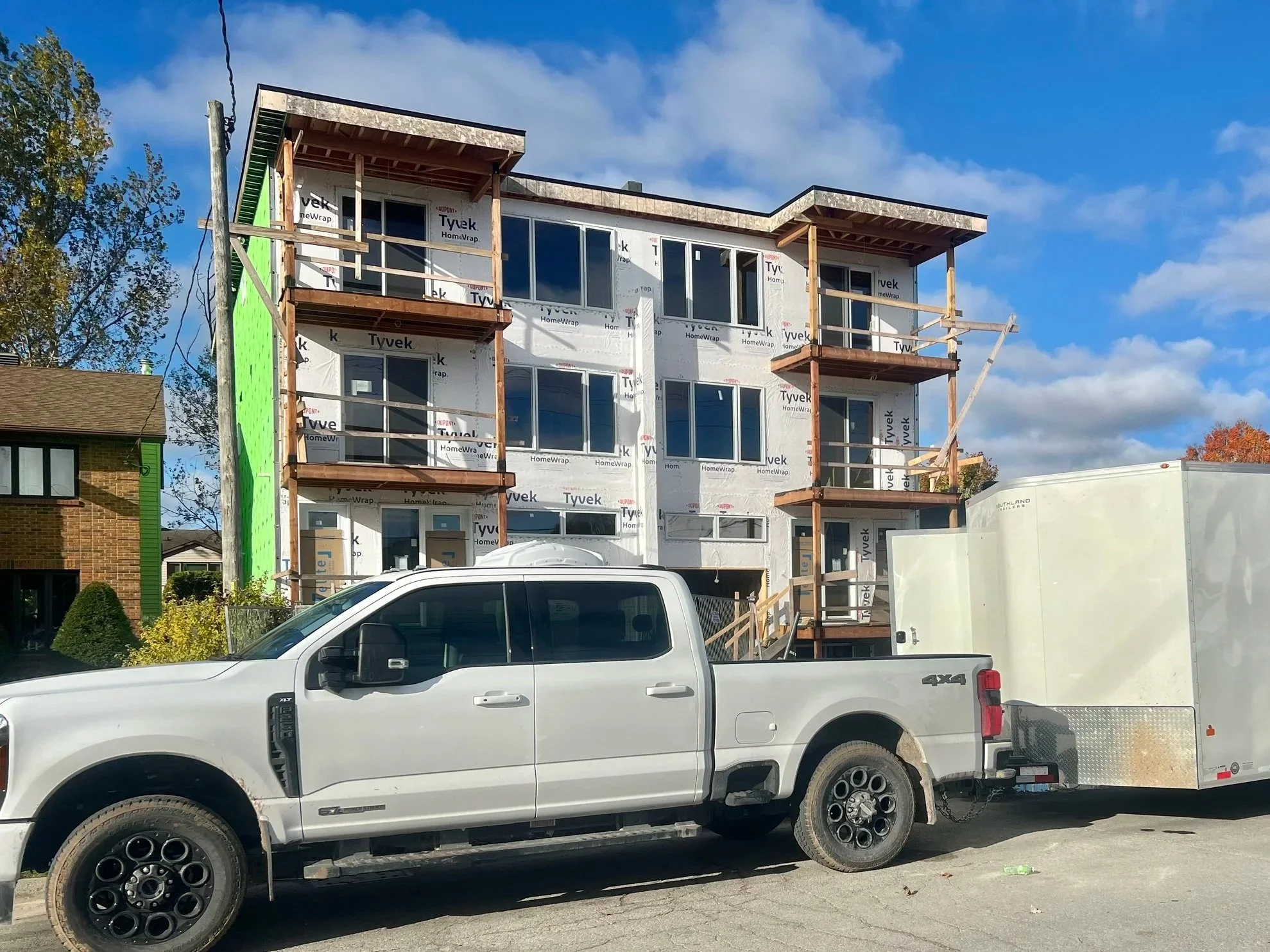 A white pickup truck parked in front of a building under construction. The building has multiple large windows and is covered with Tyvek weather barrier wrap. Wooden scaffolding surrounds the building, and it appears to be a multi-story structure. There is a green section on the left side of the building. The sky is partly cloudy with patches of blue visible.
