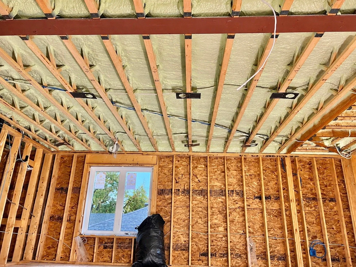 Interior view of a room under construction with exposed wooden framing, insulation, and electrical wiring, and a window showing an outdoor scene with trees and a roof.