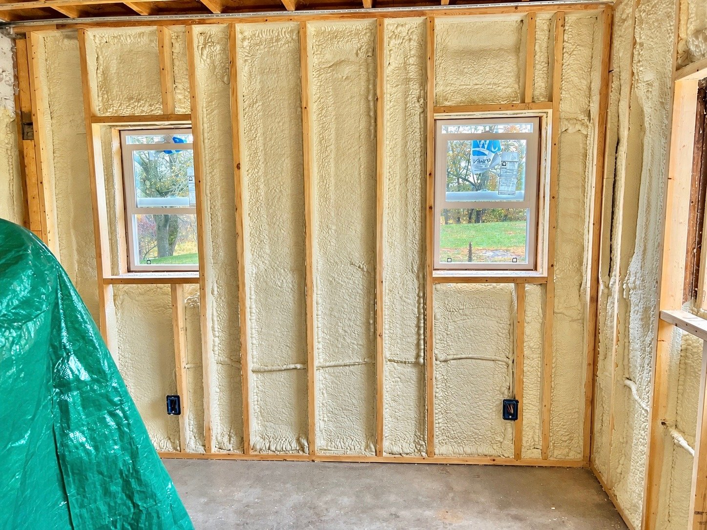 Interior of a house under construction with exposed wooden studs, spray foam insulation, and two new windows installed. There are electrical outlets and pipes visible, and part of a green tarp is on the left side.