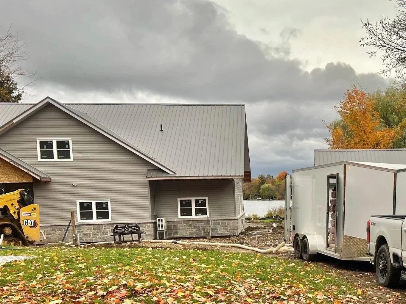Construction site with a house with grey siding and a metal roof, construction equipment, a trailer, and fallen autumn leaves on the ground.