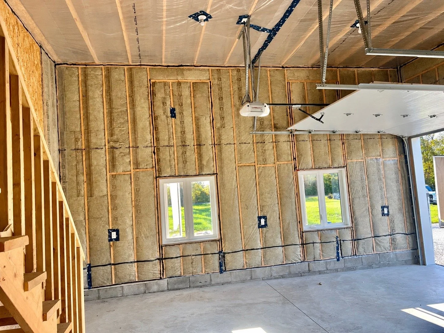 Interior of a house under construction, showing unfinished walls with insulation, electrical wiring, a garage door, and two windows. Wooden stairs are visible on the left.