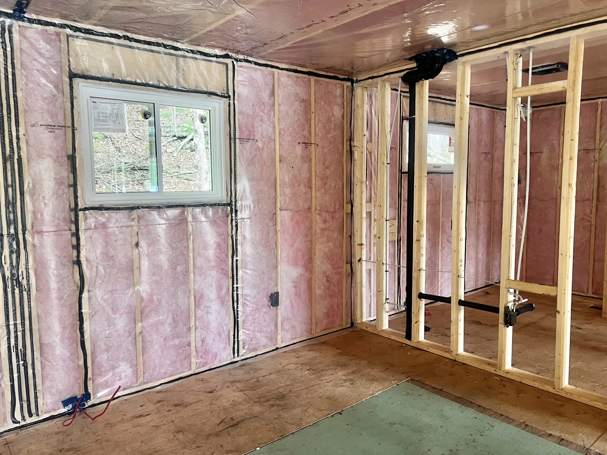 Interior of a house under construction with exposed insulation, electrical wiring, and wooden framing, with a window looking outside.