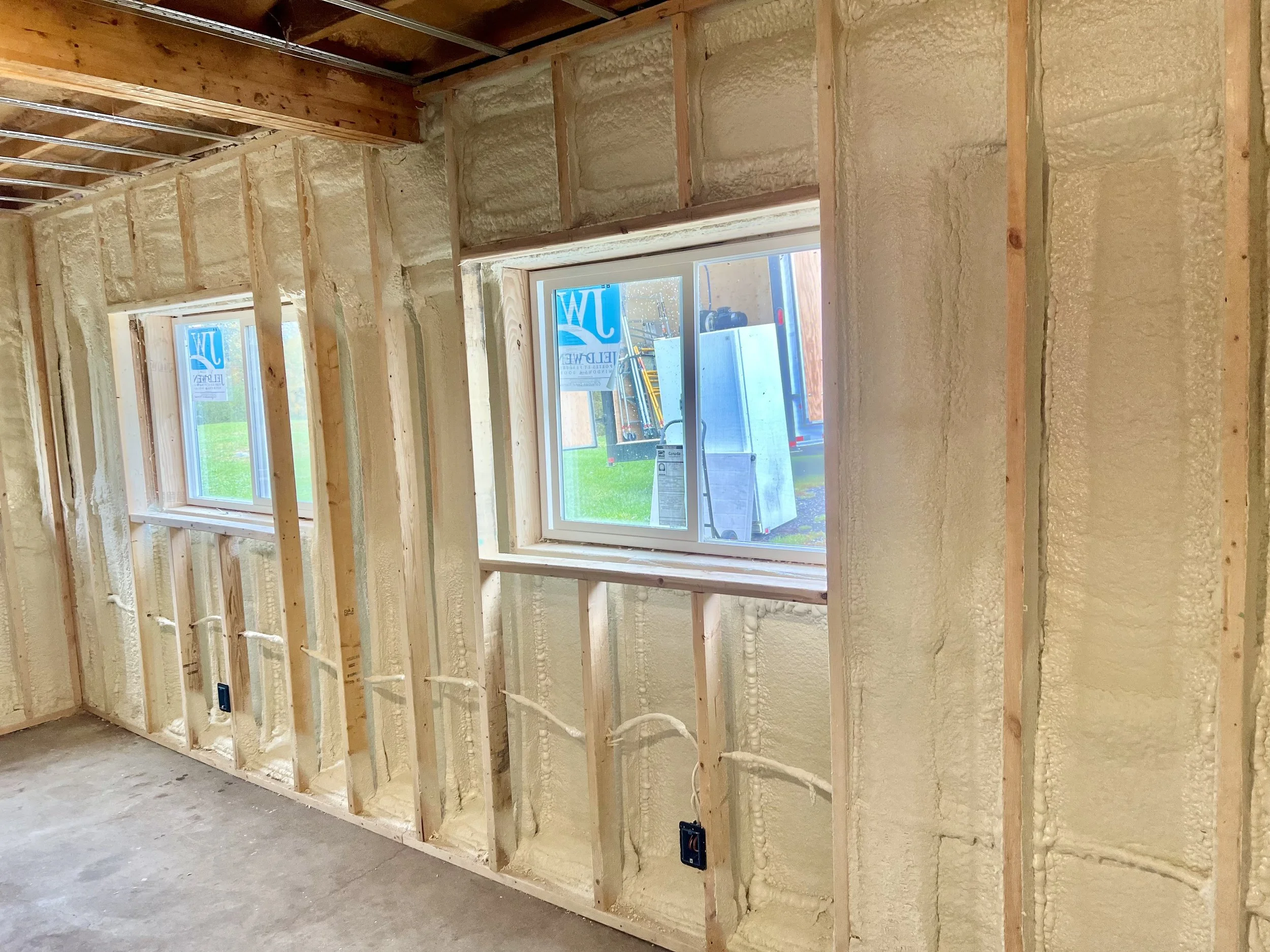Interior view of a room under construction with exposed wooden framing and spray foam insulation in the walls. Two windows are visible, letting in natural light and showing an outdoor scene with grass and a blue trailer.