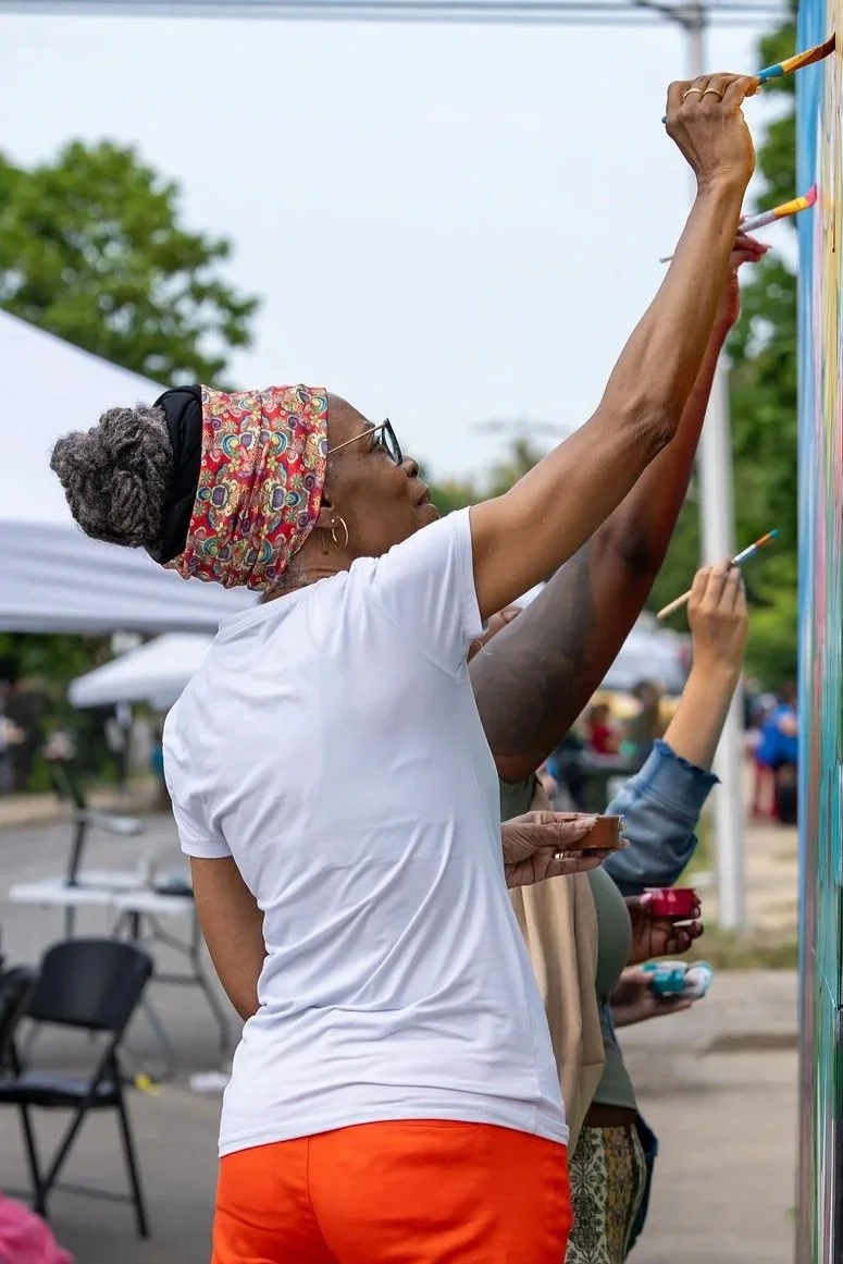Community members collaborate on painting a colorful outdoor mural at a neighborhood block party, adding bright colors to the large public mural.