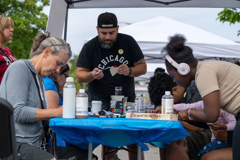 Community members of varied ages create handmade art together at a neighborhood block party, painting and decorating crafts at a table under an outdoor tent.