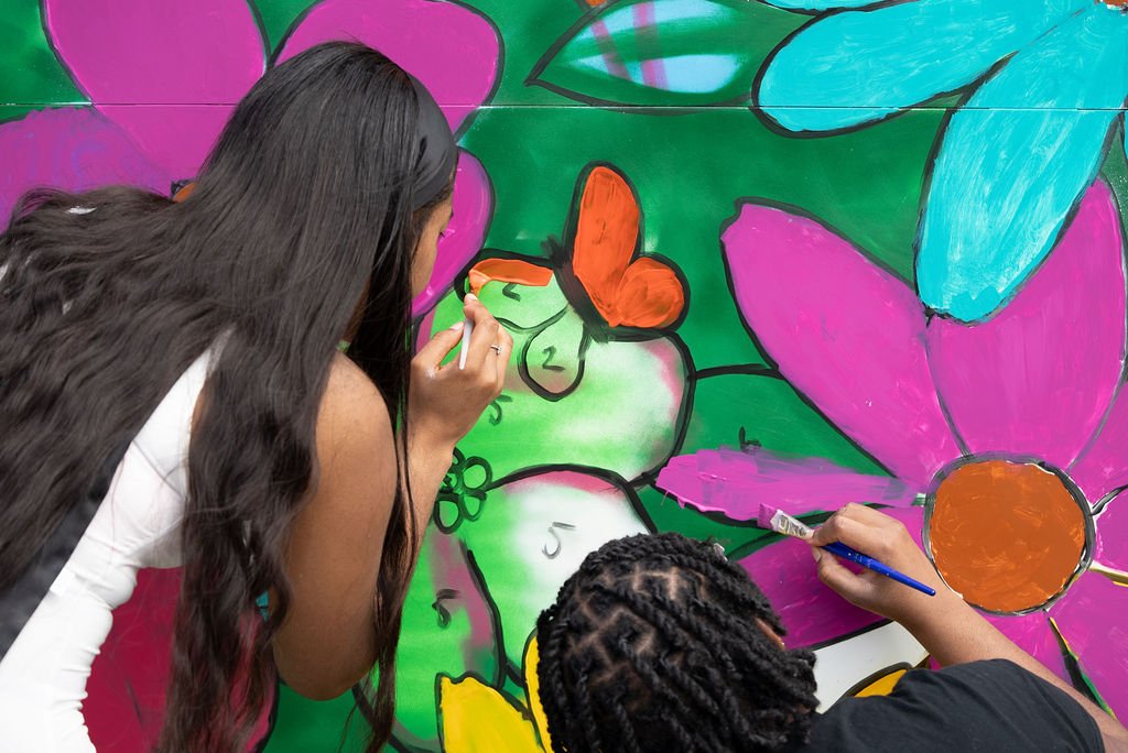Two people painting a colorful community mural featuring large flowers and butterflies during a neighborhood block party.