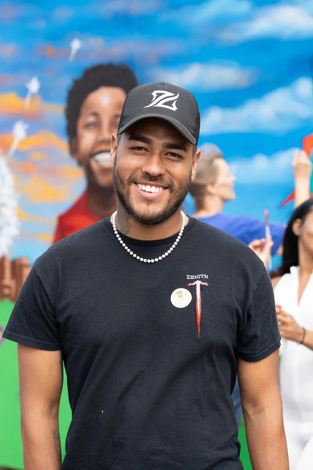 Artist Everett Reynolds stands smiling in front of a large mural being painted by community members.
