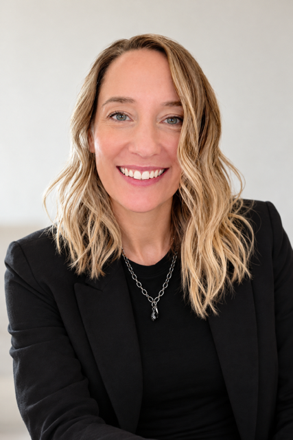 A smiling woman with shoulder-length blonde hair wearing a black blazer and necklace, sitting against a plain background.