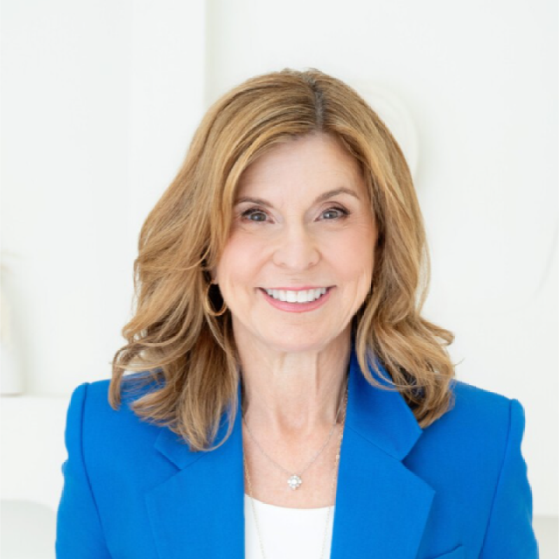 A smiling woman with shoulder-length light brown hair, wearing a blue blazer and a white top, sitting indoors with a white background.
